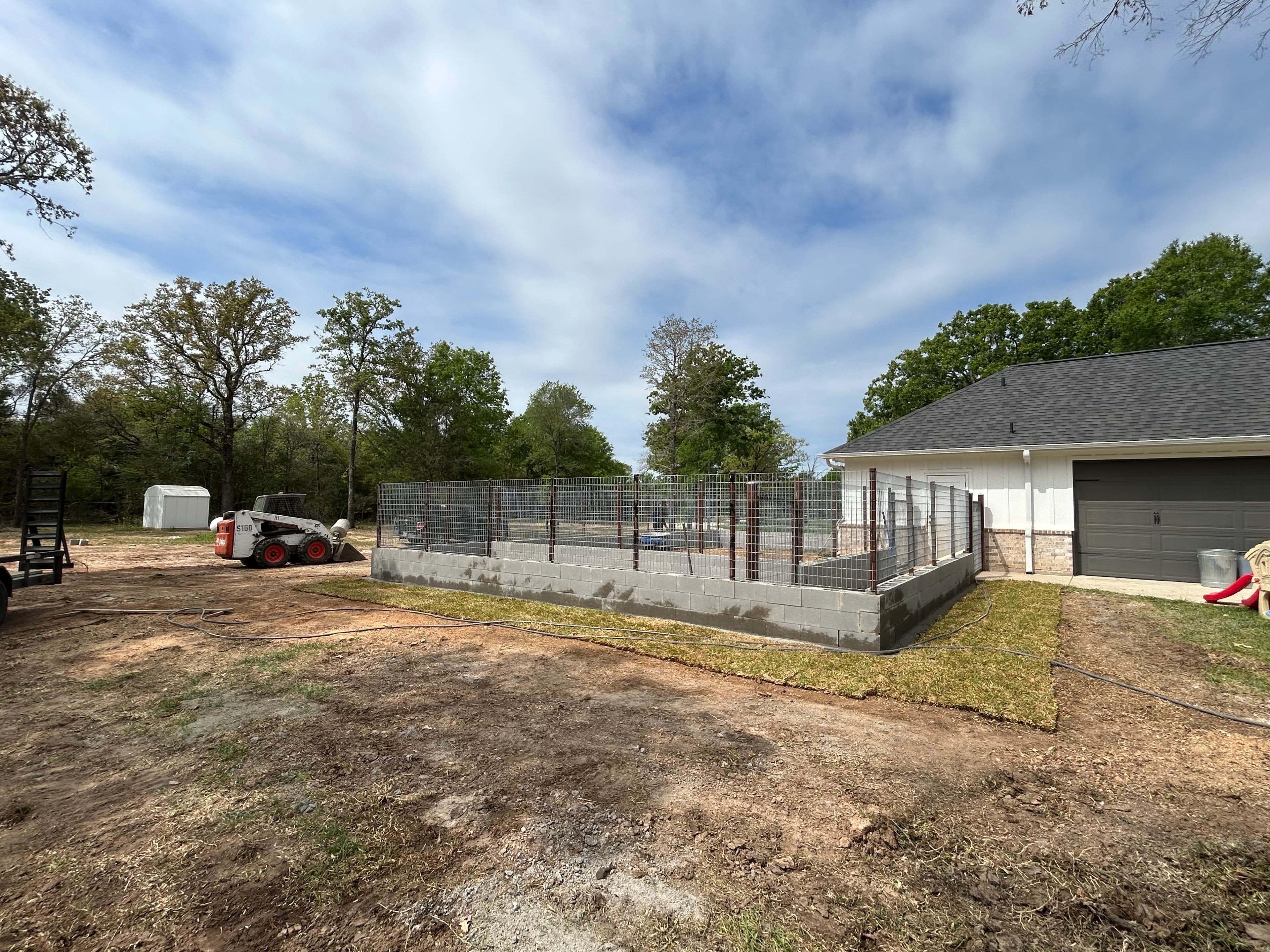 Concrete block and wire mesh enclosure under construction in a yard with a Bobcat.